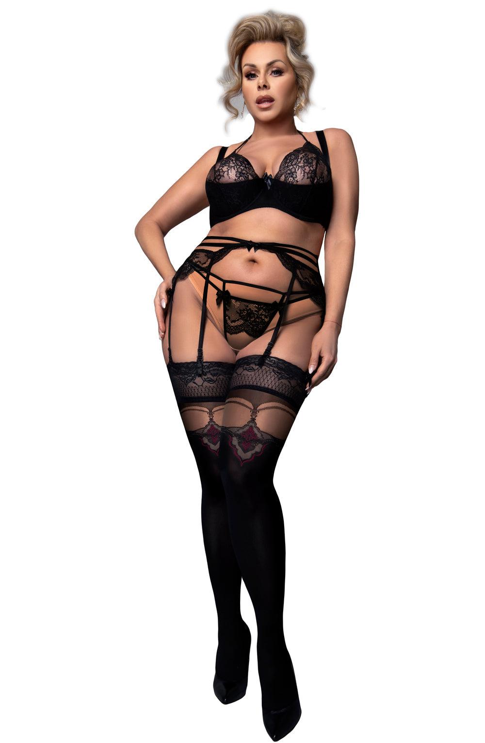 A woman stands confidently wearing a matching set of black lace lingerie, including a bra, panties with faux suspender detail, and the Ballerina 602 Black Hold Ups Plus stockings by Ballerina. She has her hair styled in loose curls and wears high heels. Her expression is assertive, and she poses with one hand on her hip.