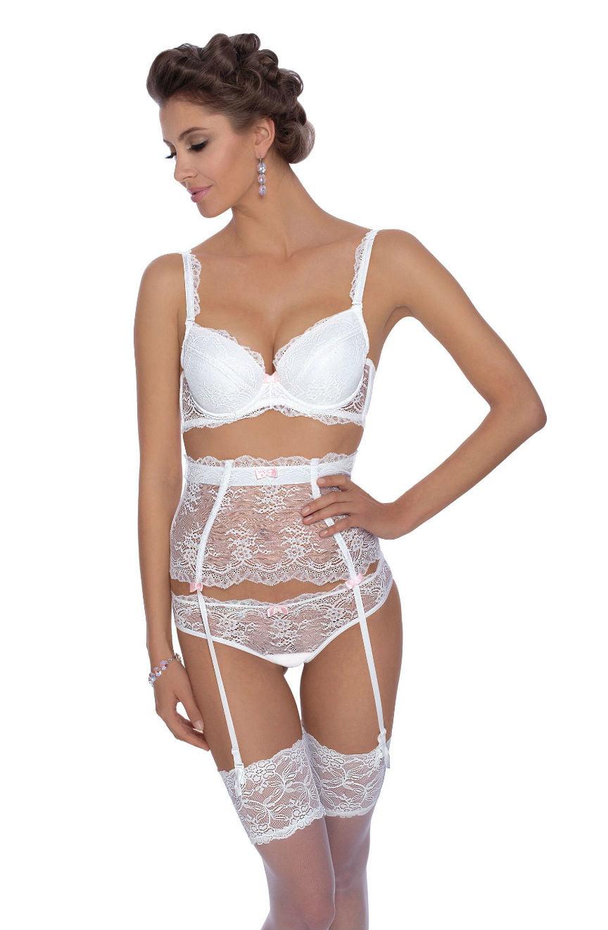 A woman stands modeling elegant white lingerie, featuring a lacy bra, high-waisted garter belt, and the matching Roza Ambre Thong White by Roza. Her hair is styled in an updo and she wears dangling earrings, facing slightly to the side with a soft smile.