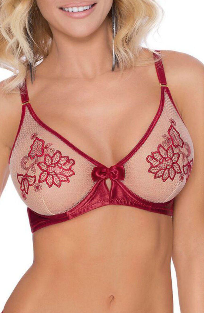 A woman with shoulder-length blonde hair, wearing the Roza Mehendi Soft Cup Bra Claret by Roza, smiles. The red and beige bra is adorned with exquisite floral embroidery that resembles Mehendi designs. It features thin straps and a delicate bow in the center.