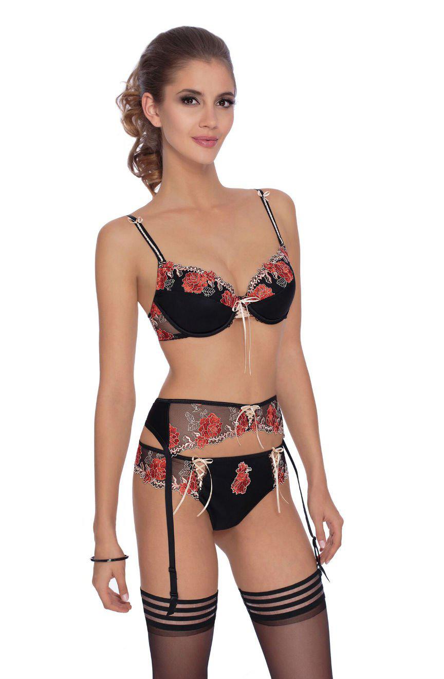 A woman with a ponytail is modeling the Roza Natali Black Bra adorned with red floral embroidery and lace-up detail. The set, from the Roza brand, includes a black bra, garter belt, and matching panties. She completes the look with black thigh-high stockings featuring horizontal stripes near the top. The background is plain white.