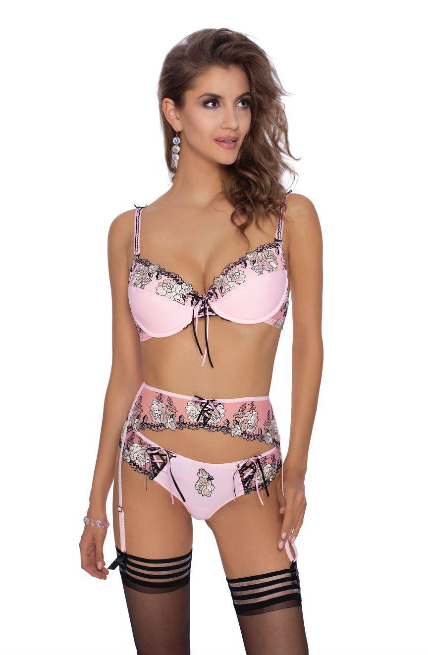 A woman is confidently modeling the Roza Natali Pink Suspender Belt lingerie set, featuring a bra, garter belt, and panties adorned with black lace accents. Her long wavy hair cascades gracefully as she wears elegant earrings. She completes her alluring look with matching thigh-high stockings that showcase exquisite floral embroidery and black bands at the top.