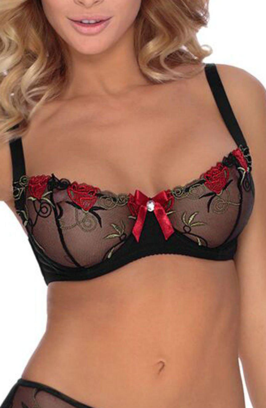 A person wearing the elegant Roza Rufina Soft Cup Bra Black with sheer cups adorned with red rose floral embroidery and green leaf details. The bra has red ribbon accents and a small central bow. The person, with wavy blonde hair, is posing against a neutral background.