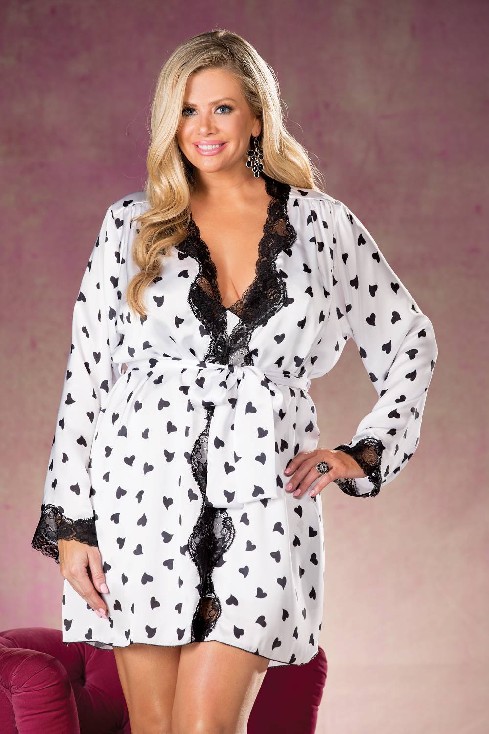 A woman with long, wavy blonde hair stands against a pink background, smiling at the camera. She wears a stunning Shirley Of Hollywood X25799 Heart Print & Lace Robe from Shirley of Hollywood, featuring black heart patterns and delicate lace trim. The robe is tied with a belt at the waist. She accessorizes with drop earrings and a ring.