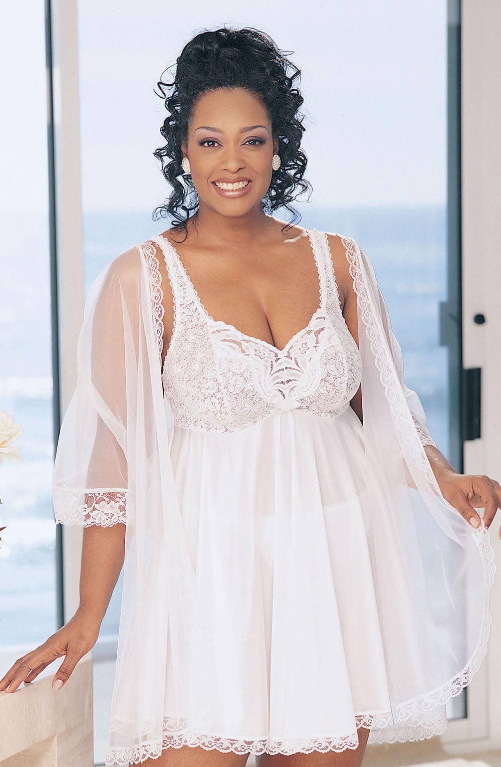 A woman is smiling and standing indoors near a large, ocean-view window. She is wearing the Shirley of Hollywood X3595 White 3 Piece Babydoll from Shirley of Hollywood, which features a white, sheer lace robe with lace trim and stretch lace cups. Her elegant hairdo showcases curls cascading down, and the natural light beautifully highlights her radiant complexion.