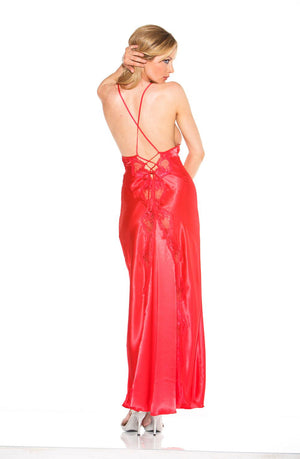 A woman stands posing with her back to the camera, wearing the Shirley Of Hollywood 20300 Red Long Gown by Shirley of Hollywood, featuring a low-cut back and crisscross tie shoulder straps. She is holding her hair with one hand while her right arm rests on her hip. She completes the look with white heeled shoes.